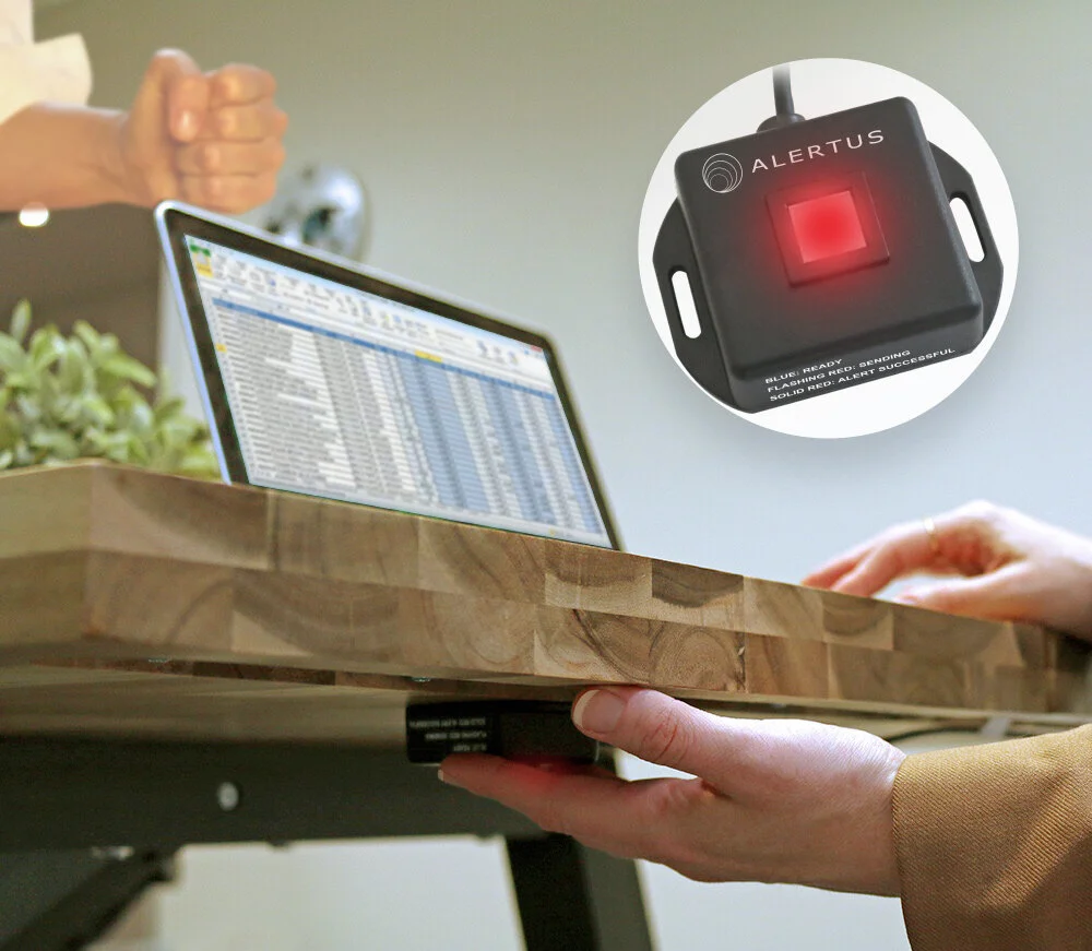 Employee hand pressing Alertus under-desk panic button in office environment with laptop and communication device