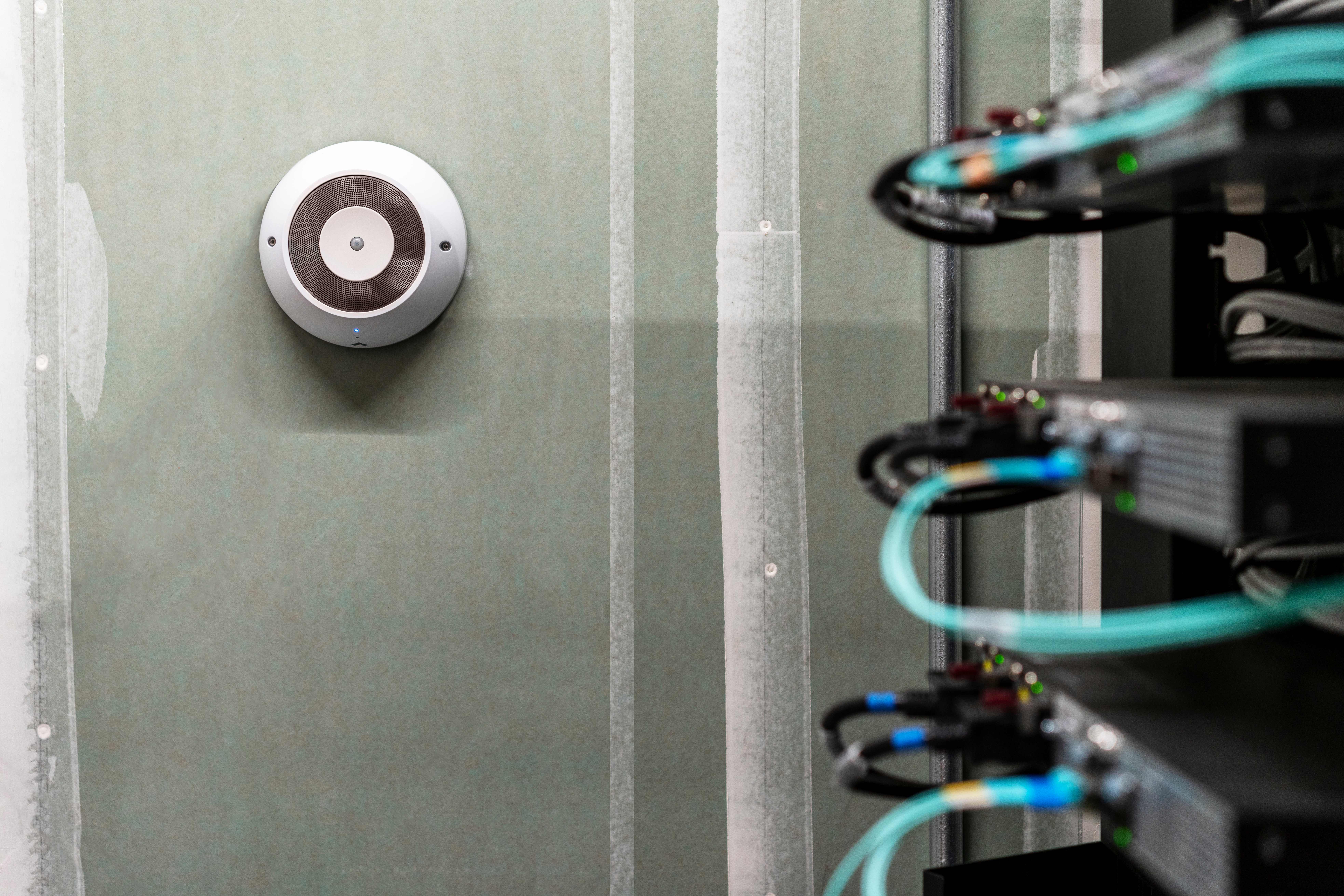 Circular environmental sensor mounted on wall in server room with network equipment and cables